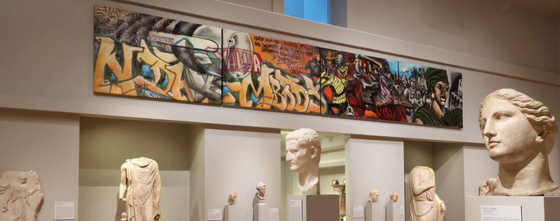 Installation view of one of the Greek and Roman Art Galleries featured a graffiti work by Docta hanging on the wall with classical sculptures on pedestals 
